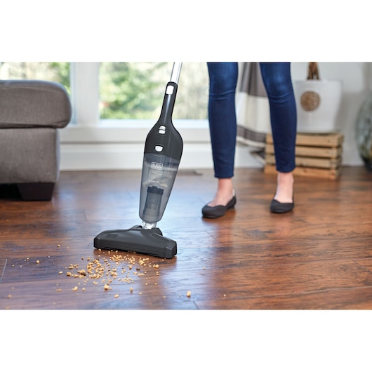 A person using a BLACK+DECKER stick vacuum cleaner to clean crumbs on a wooden floor in a living room setting.
