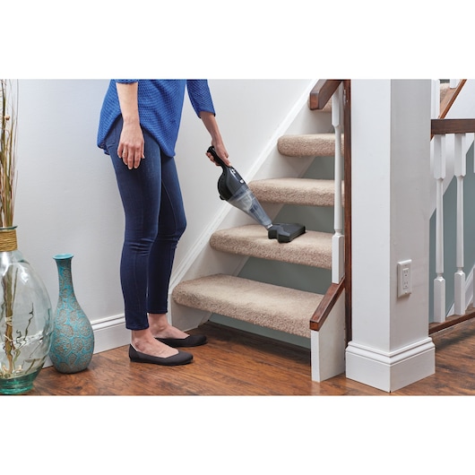 A person using a BLACK+DECKER handheld vacuum cleaner to clean carpeted stairs indoors. The person is standing on a wooden floor next to the stairs.