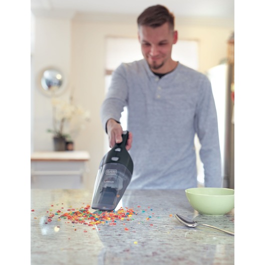 A person using a BLACK+DECKER handheld vacuum cleaner to pick up colorful cereal pieces scattered on a kitchen countertop. A green bowl and a spoon are visible on the counter.