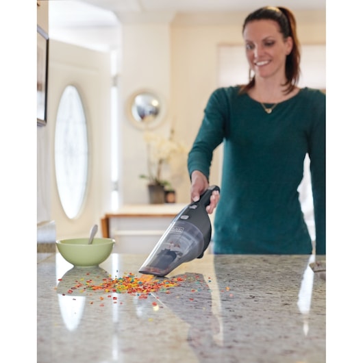 A person using a BLACK+DECKER handheld vacuum cleaner (model NSVA315J) to clean colorful cereal crumbs from a kitchen countertop. A green bowl with a spoon is placed nearby.