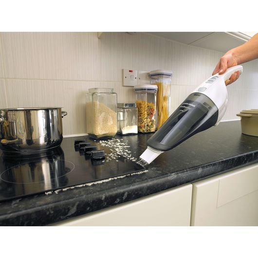 A BLACK+DECKER handheld vacuum cleaner being used to clean spilled grains from a kitchen countertop near a stove, with jars of food in the background.