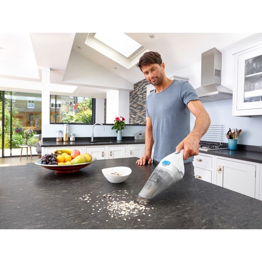 A person using a BLACK+DECKER handheld vacuum cleaner to clean spills of cereal on a kitchen countertop. There is a fruit bowl and a white bowl nearby in a modern kitchen with large windows.