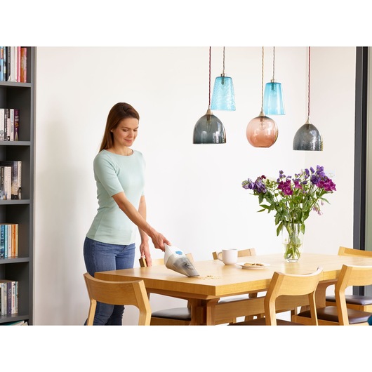 A person using a BLACK+DECKER handheld vacuum cleaner to clean crumbs from a wooden dining table in a modern home setting. There are flowers, cups, and plates on the table, and pendant lights hanging overhead.