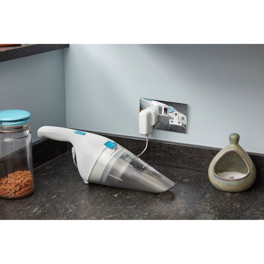 A handheld cordless vacuum cleaner with white and blue accents, shown charging on a kitchen countertop next to a glass jar of pasta and a ceramic container.