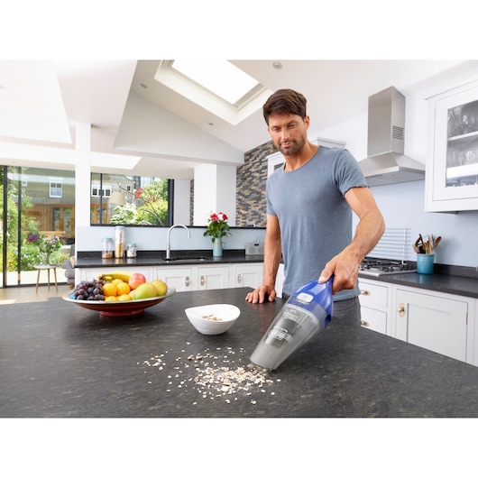 A person using a BLACK+DECKER handheld vacuum cleaner (model NVC115WA) to clean spilled oats on a kitchen countertop. A bowl and a fruit platter are also on the counter in a modern kitchen setting.