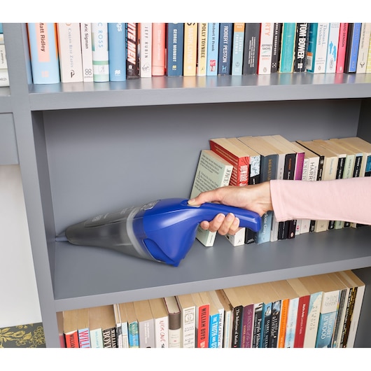 A person is using a blue BLACK+DECKER handheld vacuum cleaner to clean dust from a gray bookshelf filled with books.