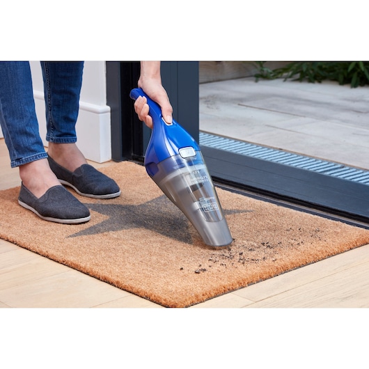 A person using a handheld BLACK+DECKER vacuum cleaner to remove dirt from a doormat next to a doorway.