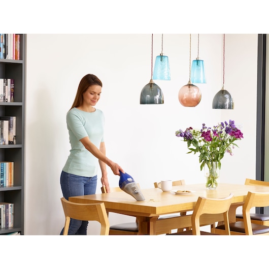A person is using a BLACK+DECKER handheld vacuum (model NVC115WA) to clean crumbs off a wooden dining table. The table has chairs around it, a vase with purple flowers, and a cup. Modern pendant lights hang above, and a bookshelf is visible in the background.