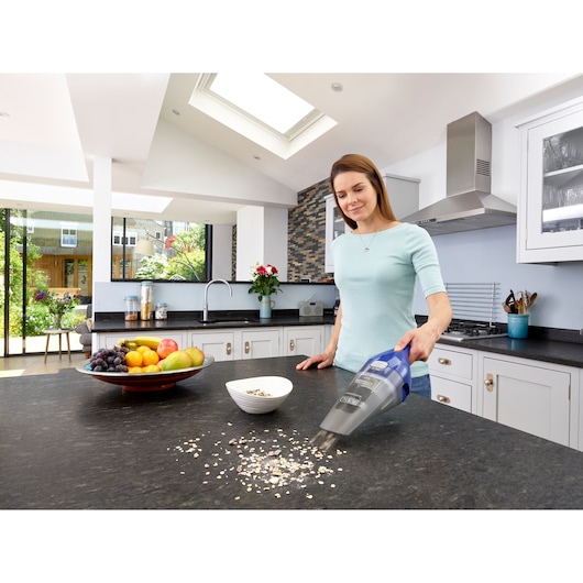 A person using a BLACK+DECKER NVC115WA handheld vacuum cleaner to clean food debris from a kitchen countertop. A bowl of fruit and a small white bowl are also visible on the counter in a modern kitchen setting.