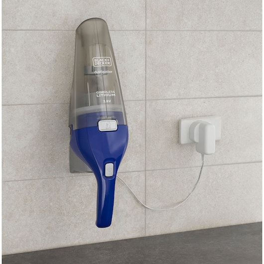 A BLACK+DECKER cordless handheld vacuum cleaner (dustbuster) is mounted on a tiled wall, plugged in and charging. The vacuum has a blue and grey design and is labeled 'Cordless Lithium 3.6V.'