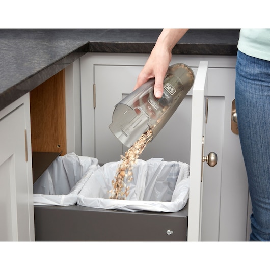 A person empties the dirt container from a Black+Decker handheld vacuum into a trash bin with two sections, inside a kitchen cabinet.