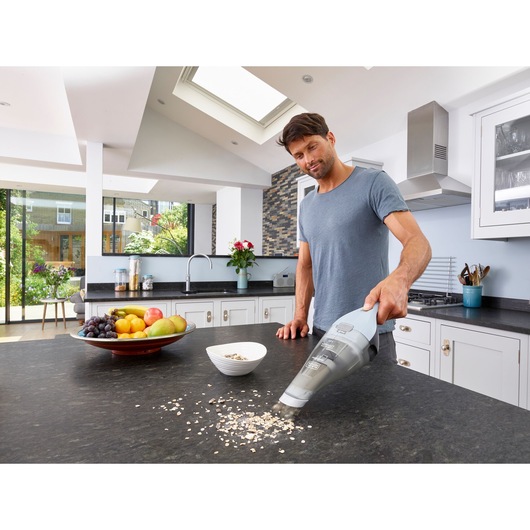 A person using a BLACK+DECKER handheld vacuum cleaner (model NVC215W) to clean spilled oats on a kitchen countertop. A bowl of fruit and a white bowl are also on the counter in a modern kitchen setting.