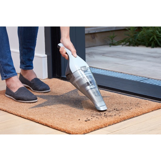 A person uses a BLACK+DECKER handheld vacuum cleaner to clean dirt from a doormat at a doorway.