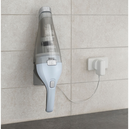 A BLACK+DECKER cordless handheld vacuum cleaner model NVC215W is mounted on a wall charging station, connected to a power outlet. The device is grey and light blue, and is shown in a modern indoor setting with tiled walls.