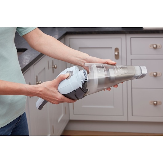 A person is detaching the dust container from a BLACK+DECKER handheld cordless vacuum cleaner in a modern kitchen setting.