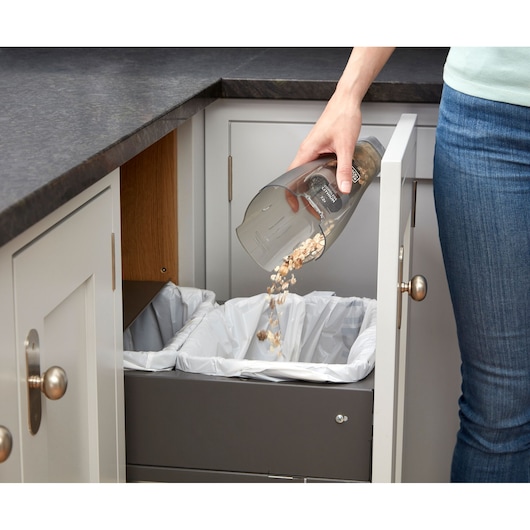 A person empties debris from a handheld BLACK+DECKER vacuum cleaner into a pull-out kitchen trash bin lined with white plastic bags.