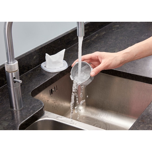 A person rinsing a round vacuum filter under running water in a kitchen sink. The filter is held under a modern faucet, and another filter part is placed on the countertop nearby.