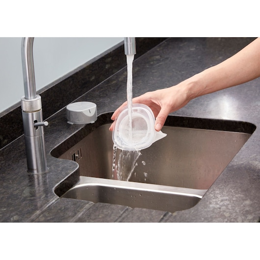 A person is rinsing a removable filter under running water at a kitchen sink. The filter appears to belong to a household appliance, possibly a vacuum cleaner.