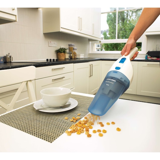 A person using a BLUE BLACK+DECKER handheld vacuum cleaner to pick up cereal from a white table in a modern kitchen. The vacuum is held over the spilled cereal next to a white bowl and plate.