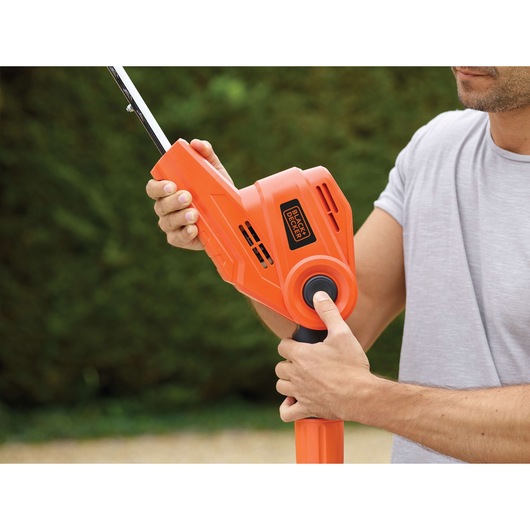 A person operating a BLACK+DECKER orange hedge trimmer outdoors, holding and adjusting the tool's handle.
