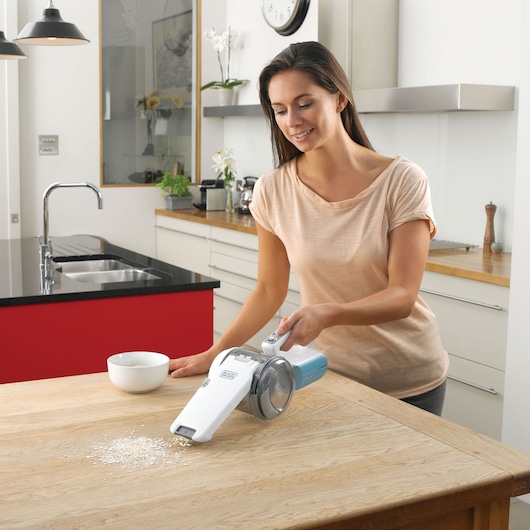 A person using a BLACK+DECKER PV1020L handheld vacuum cleaner to remove spilled oats from a wooden kitchen table. The kitchen is modern with light cabinetry, a bowl on the table, and a clock on the wall.