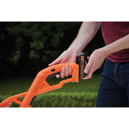 A person is attaching an 18V lithium battery to the handle of an orange BLACK+DECKER power tool, with greenery in the background.