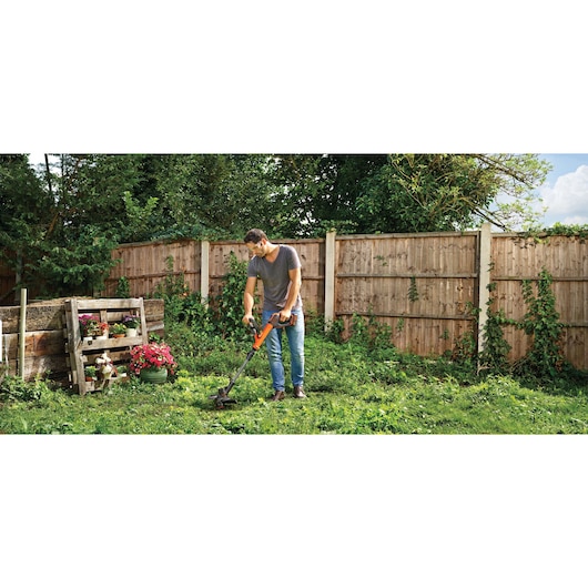 A person using a BLACK+DECKER cordless grass trimmer in a garden, surrounded by a wooden fence and greenery. There are flowers planted in containers on the left side.