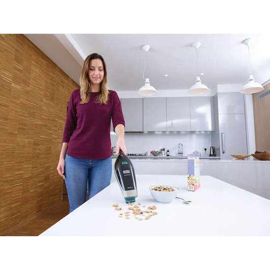 A person using a BLACK+DECKER handheld vacuum cleaner to pick up cereal pieces from a white kitchen table. There is a bowl of cereal, a spoon, and a cereal box on the table in a modern kitchen setting.
