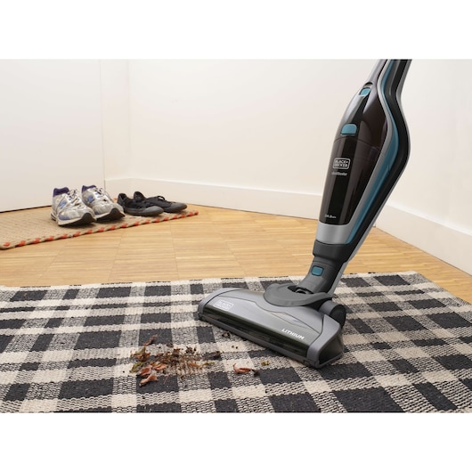 A BLACK+DECKER SVA420B cordless stick vacuum cleaner is being used to clean dirt and leaves from a checkered rug in a home, with shoes placed on a mat near the wall in the background.