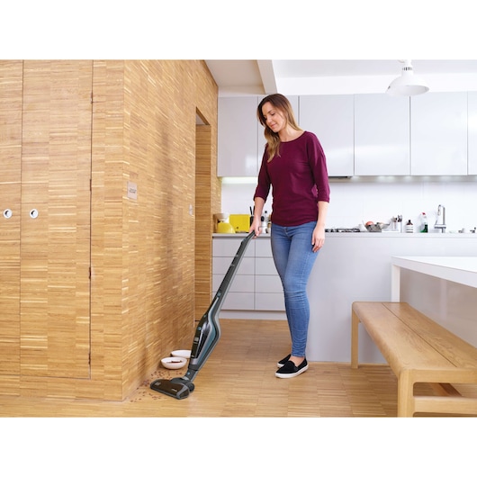 A person is using a BLACK+DECKER cordless stick vacuum to clean spilled food on a wooden kitchen floor beside a pet bowl. The kitchen has light-colored cabinets and a wooden bench.