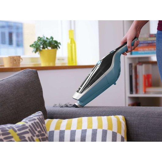 A person uses a BLACK+DECKER handheld vacuum cleaner to clean a grey sofa, with decorative pillows in the foreground. The scene is in a modern, well-lit living room with a yellow plant pot and books visible in the background.