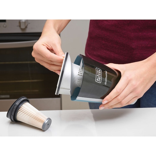 A person is cleaning or removing the filter from a BLACK+DECKER dustbuster handheld vacuum. The vacuum and its removable filter are placed on a white counter in a kitchen setting.