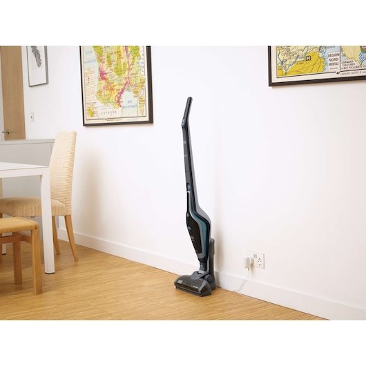A BLACK+DECKER upright cordless vacuum cleaner, model SVA420B, standing on a wooden floor and charging via a wall outlet in a modern room with maps on the wall and a dining table with chairs.