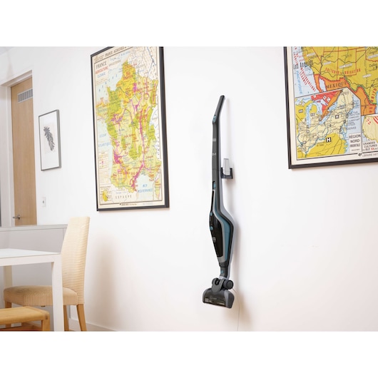 A BLACK+DECKER cordless stick vacuum cleaner (model SVA420B) mounted on a wall in a modern room. The room features two framed map posters and a chair next to a table.