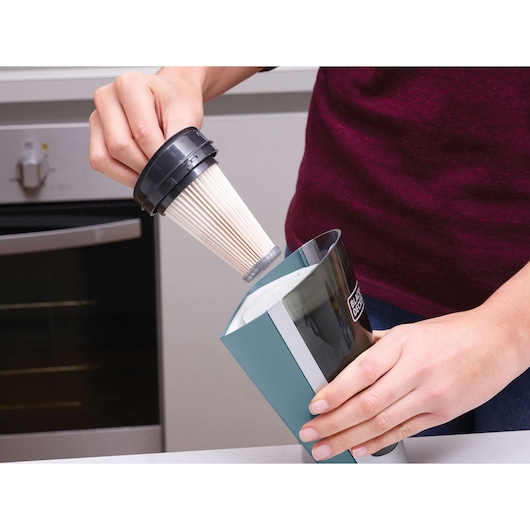 A person is removing a filter from a BLACK+DECKER handheld vacuum cleaner while standing in a kitchen.