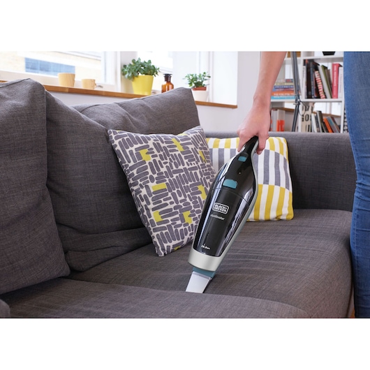 A hand-held BLACK+DECKER vacuum cleaner is being used to clean a gray fabric sofa with patterned cushions in a living room.