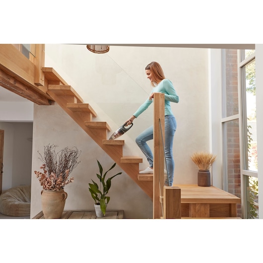 A person using a BLACK+DECKER handheld vacuum cleaner to clean a wooden staircase inside a modern home. Decorative plants and vases are positioned at the base of the stairs, and large windows let in natural light.
