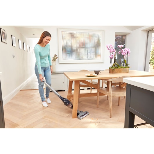A person using a BLACK+DECKER cordless stick vacuum cleaner (model SVB520JW) to clean crumbs from a light wooden floor near a dining table with pink orchids and a cup on top. The scene is set in a bright and modern dining area.