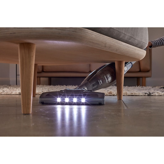 A BLACK+DECKER cordless vacuum cleaner with LED lights is shown cleaning under a sofa on a hard floor.