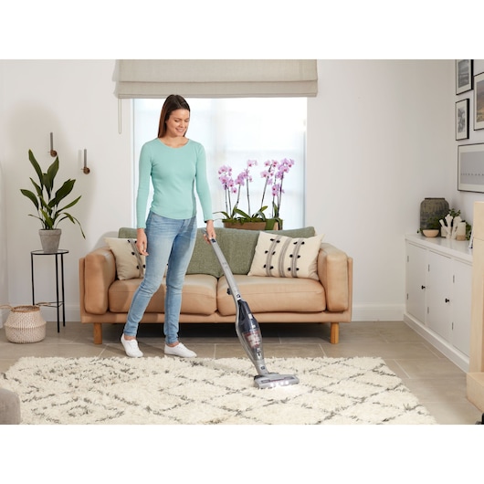A person is vacuuming a light-colored rug in a modern living room with a BLACK+DECKER upright vacuum cleaner model SVB520JW. The room has a beige sofa, green and cream cushions, pink orchids on a window sill, and various decorative items.