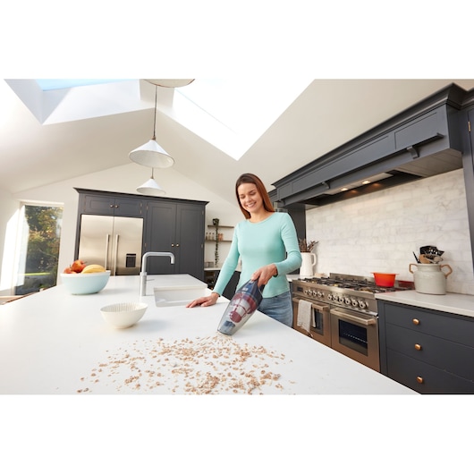 A person using a BLACK+DECKER handheld vacuum cleaner (model SVB520JW) to clean crumbs from a kitchen countertop in a modern kitchen.