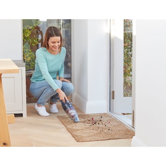 A person is using a BLACK+DECKER handheld vacuum cleaner to clean dirt and debris from a woven mat near a door inside a bright room.