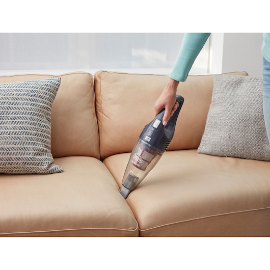 A person using a BLACK+DECKER handheld vacuum cleaner to clean the crevice between the cushions of a light brown sofa.