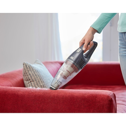 A BLACK+DECKER handheld vacuum cleaner is being used to clean a red sofa, with a patterned cushion visible in the background.