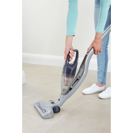 A person is using a BLACK+DECKER cordless stick vacuum cleaner on a light-colored tiled floor. The vacuum has a detachable handheld unit and is being operated by someone wearing light blue jeans, a turquoise long-sleeve top, and white shoes.