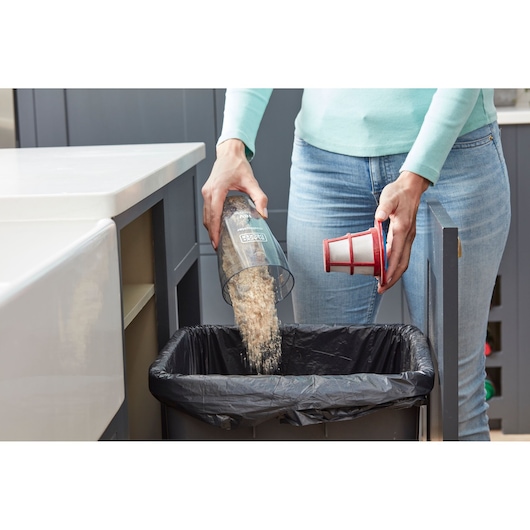 A person empties debris from a BLACK+DECKER vacuum cleaner dust container into a kitchen trash bin. The person is holding the vacuum's filter in one hand and pouring the contents out with the other.