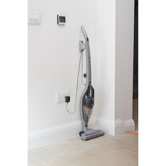 A cordless stick vacuum cleaner, plugged in and charging at a wall outlet, positioned upright against a white wall in a modern home interior.