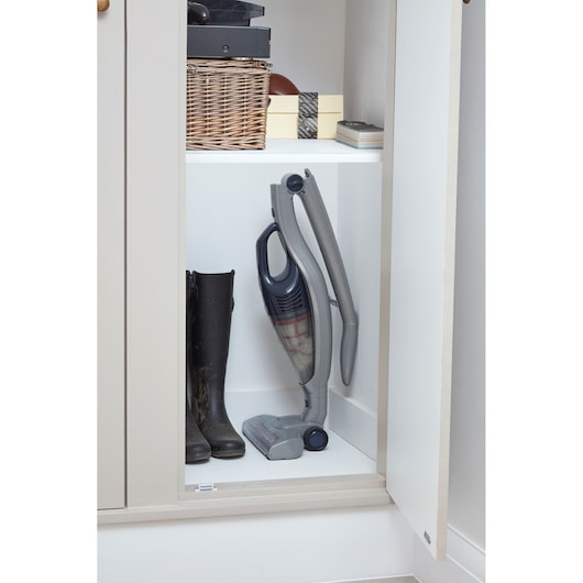 A cordless stick vacuum cleaner stored upright in a cabinet next to a pair of muddy black boots. Above, there are baskets, boxes, and other storage items on the shelf.