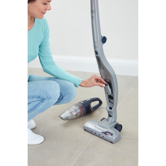 A person kneeling on the floor and handling a BLACK+DECKER 2-in-1 cordless stick vacuum cleaner, with the handheld vacuum attachment placed on the floor next to the main unit.