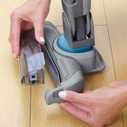 Close-up of hands removing the brush roll from a BLACK+DECKER vacuum cleaner on a wooden floor.